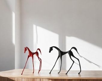 May include: Two minimalist horse sculptures. One is a vibrant red, the other is black. Both have elongated legs and arched necks, giving them a modern, artistic look. They stand on a wooden surface against a white wall, creating a striking contrast.