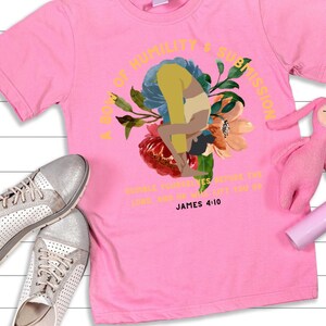 Puede incluir: Camiseta rosa con un diseño floral y el texto "A Boy of Humility & Submission". El diseño incluye una figura en una postura de yoga. El texto "Humble yourselves before the Lord, and He will lift you up. James 4:10" también está impreso en la camiseta.