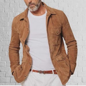 May include: A brown suede jacket with four front pockets and button closures. The jacket is worn over a white t-shirt and paired with white pants and a brown belt. The jacket has a classic, tailored design.