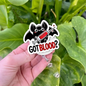 May include: A badge reel with a cartoon bat holding a syringe filled with red liquid. The bat is black with white accents and has a smiling face. The text "GOT BLOOD?" is in white with a red border.