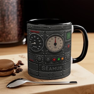 May include: Black coffee mug with a steampunk-inspired design. The mug features a panel with gauges, dials, and the name "SÉAMUS". The mug has a black interior and handle, and is sitting on a wooden surface.