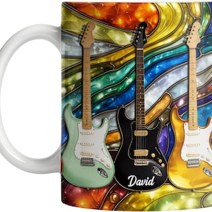 以下が含まれることがあります： 白のセラミックマグ。ミントグリーン、黒、ゴールドのエレキギター3本が描かれた、カラフルなステンドグラスのデザイン。黒いギターには「David」とプリントされています。マグには白いハンドルが付いています。