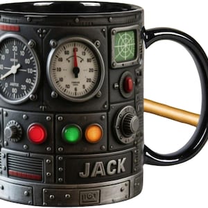 May include: Black ceramic mug with a steampunk design. The mug is decorated with gauges, dials, and lights, and the name "JACK" is printed on the front. The handle is black, and a gold-coloured object rests on the mug.