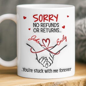 Puede incluir: Taza de cerámica blanca con el texto "SORRY NO REFUNDS OR RETURNS..." en rojo. Debajo del texto están los nombres "John" y "Emily" en escritura roja, con un símbolo de corazón. La taza también presenta un dibujo de dos manos tomadas, con el texto "...You're stuck with me forever."