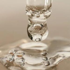 May include: Close-up of a clear glass dropper dispensing a liquid, forming a large droplet. The droplet is about to fall onto a surface, with smaller droplets already present. The liquid appears transparent with visible bubbles.