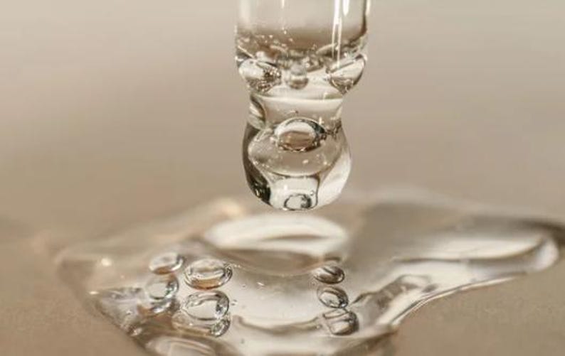 May include: Close-up of a clear glass dropper dispensing a liquid, forming a large droplet. The droplet is about to fall onto a surface, with smaller droplets already present. The liquid appears transparent with visible bubbles.