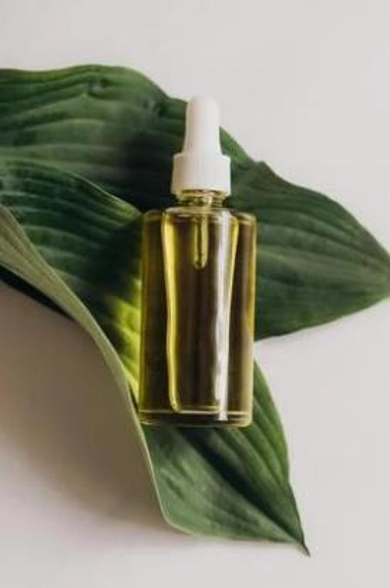 May include: A clear glass dropper bottle filled with a yellow liquid, set against a large green leaf. The bottle has a white dropper top. The background is a soft white.