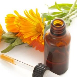 May include: A brown glass essential oil bottle with a dropper, next to bright orange calendula flowers and green leaves. The dropper is clear with a yellow liquid inside. The bottle has a black cap and white tip.