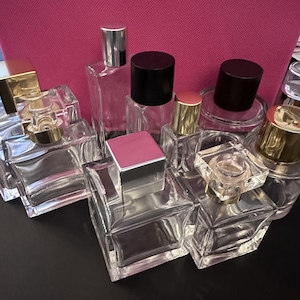 May include: An assortment of clear glass perfume bottles with different cap styles. Some caps are silver, black, or gold, and some have a pink or gold accent. The bottles are displayed on a dark surface.