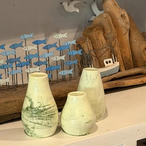 May include: Three small, pale green ceramic vases with a textured finish. The vases are arranged on a white shelf, with a driftwood and fish art piece in the background. The vases have a coastal theme.