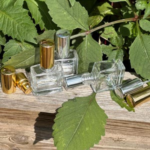 May include: Several clear glass perfume bottles with gold and silver caps are displayed. The rectangular bottles are empty, with the caps placed nearby. The scene is set on a wooden surface, with green leaves in the background.