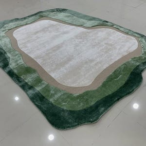 Può includere: Un tappeto morbido, di forma irregolare con un design a strati. Il tappeto presenta un'area centrale bianco sporco, circondata da fasce beige, verde chiaro e verde scuro. Dimensioni: circa 152 cm per 102 cm.