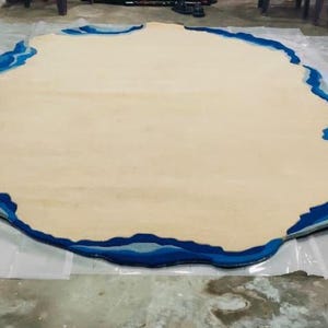 May include: A large, irregularly shaped rug with a light beige centre and a border of varying shades of blue. The rug's design resembles a coastline or island, adding a unique, artistic touch to any room. The rug is placed on a clear plastic sheet.