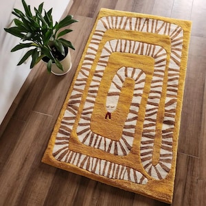 Op de afbeelding: Mosterdgele rechthoekige vloerkleed met een gestileerd slangenontwerp. De slang is afgebeeld met witte en bruine strepen en een kleine rode tong. Het vloerkleed ligt op een houten vloer en een plant in een pot is zichtbaar op de achtergrond.