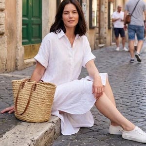 Puede incluir: Una mujer con un vestido de lino blanco y zapatillas blancas está sentada en un bordillo de piedra. Sostiene un bolso tote de paja tejida con asas de cuero marrón. El fondo muestra una calle europea con edificios y peatones.