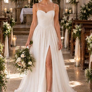 May include: A floor-length white wedding dress with thin spaghetti straps, a sweetheart neckline, and a high side slit in the skirt. The dress features a fitted bodice and a flowing, pleated skirt, paired with a sheer veil and a floral bouquet.