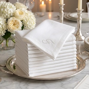 May include: A stack of white linen napkins with a gray embroidered initial on the top napkin. The napkins are neatly arranged on a silver serving tray. A bouquet of white flowers and candles are in the background.