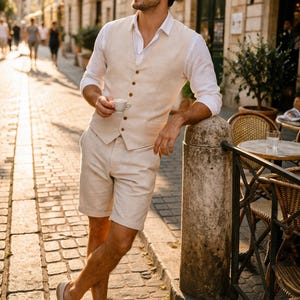 Può includere: Un uomo con gilet, pantaloncini e camicia bianca di lino beige, tiene in mano una tazzina. Indossa occhiali da sole e scarpe beige. L'ambientazione è un caffè all'aperto con strade acciottolate e un'insegna "CAFFE".