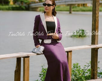 Può includere: Una donna indossa un tailleur gonna viola con un blazer con revers bianco, top nero e tacchi neri. Indossa occhiali da sole e tiene in mano una pochette argentata. Lo sfondo comprende acqua ed edifici.