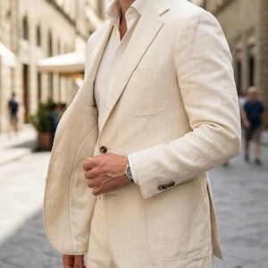 Pode incluir: Um fato de linho bege claro, composto por um blazer e calças. O blazer tem lapelas entalhadas, um bolso no peito e dois bolsos com aba. O fato é usado com uma camisa clara. O conjunto é completado com óculos de sol e um relógio de pulso.