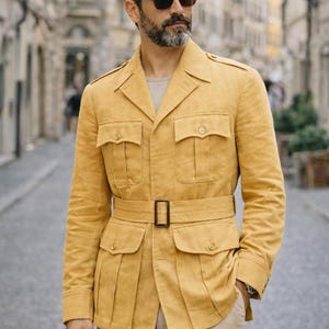 May include: Mustard yellow safari jacket with four front pockets, a belted waist, and a notched collar. The jacket is paired with khaki pants. The jacket has a classic, tailored fit and is made of a textured fabric.