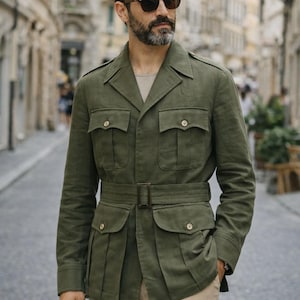 May include: An olive green safari jacket with multiple pockets and a matching belt. The jacket features a classic collar and button fastenings. The model is wearing sunglasses and beige trousers. The jacket is made of a lightweight material.