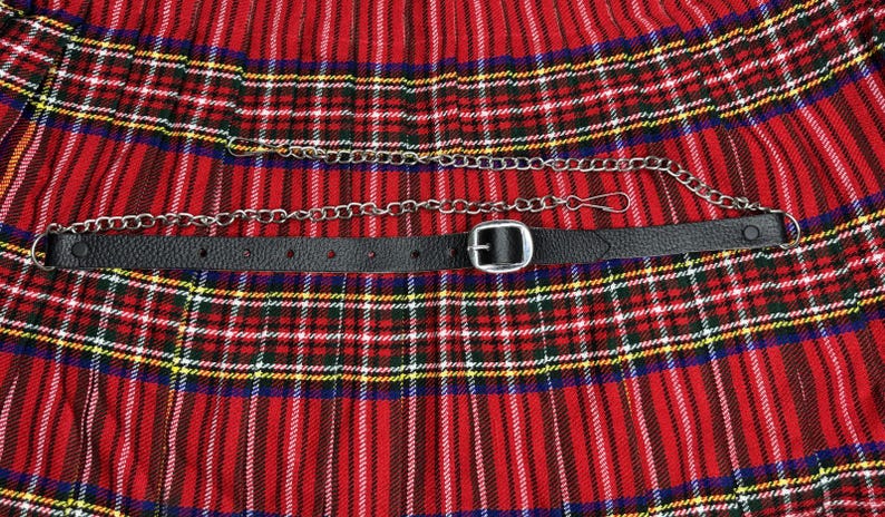 May include: A black leather belt with a silver buckle and a silver chain, set against a red, green, and yellow plaid fabric. The belt has multiple holes for adjustment. The chain is attached to the belt.