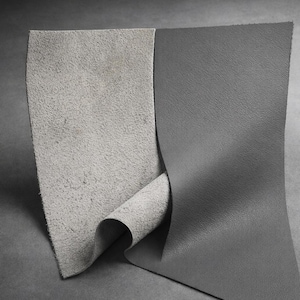 May include: A close-up of two pieces of gray leather. One piece has a rough, suede-like texture, while the other is smooth with a subtle grain. The edges of the leather are visible, showing the different textures.