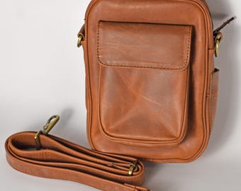 May include: A brown leather crossbody bag with a front pocket and a long, adjustable strap. The bag has a rectangular shape with rounded corners and a zipper closure. The strap and hardware are a brass color.