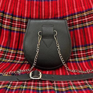 May include: A black leather sporran with a silver chain and belt, set against a red and black plaid background. The sporran has a rounded bottom and a flap closure. The belt has a silver buckle.
