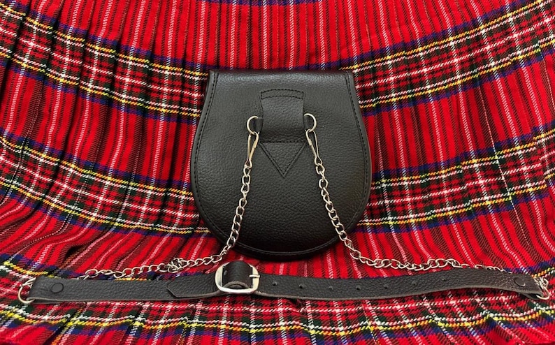 May include: A black leather sporran with a silver chain and belt, set against a red and black plaid background. The sporran has a rounded bottom and a flap closure. The belt has a silver buckle.