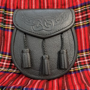May include: Black leather sporran with Celtic knotwork design on the flap. The sporran has three decorative tassels hanging from the bottom. The sporran is set against a red and black plaid background.