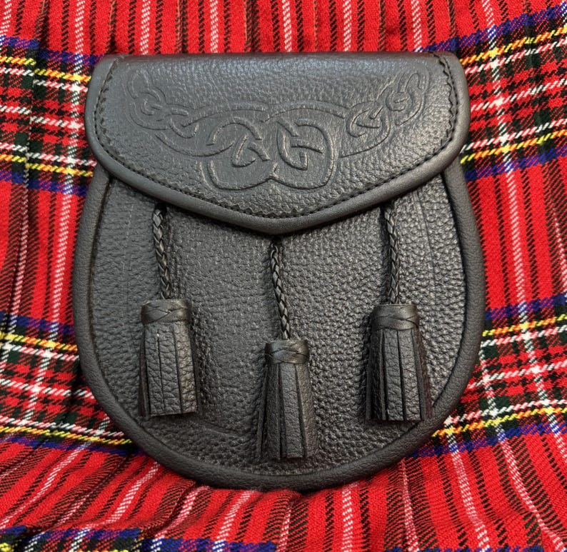 May include: Black leather sporran with Celtic knotwork design on the flap. The sporran has three decorative tassels hanging from the bottom. The sporran is set against a red and black plaid background.