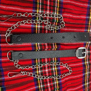 May include: Two black leather kilt straps with silver-colored chains and clasps. One strap has a silver buckle. The straps are on a red, black, and yellow plaid background.