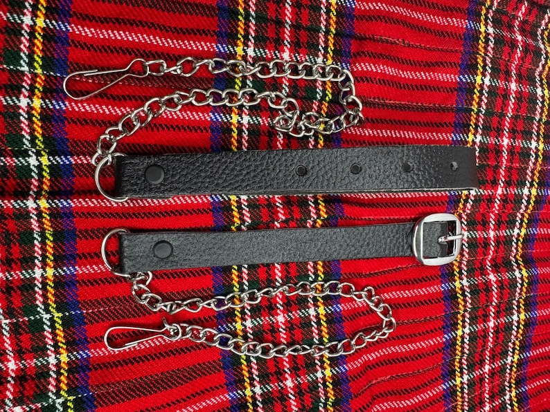 May include: Two black leather kilt straps with silver-colored chains and clasps. One strap has a silver buckle. The straps are on a red, black, and yellow plaid background.