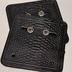 Könnte beinhalten: Zwei schwarze, quadratische Ausweishalter aus Kunstleder mit strukturierten Oberflächen und Druckknöpfen. Die Halter haben eine rechteckige Form mit abgerundeten Ecken und einem dekorativen Rand. Die Druckknöpfe sind silberfarben.