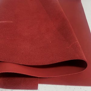 May include: A sheet of red leather, partially rolled, revealing a textured suede-like surface on one side and a smooth, flat surface on the other. The leather is a deep, rich shade of red.