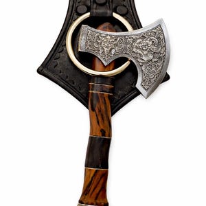 May include: An ornate axe with a silver blade featuring detailed engravings of mythical creatures. The wooden handle has a striped pattern and is accented with a decorative silver base. The axe is attached to a black leather sheath with a gold ring.