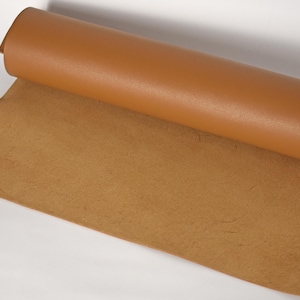 May include: A roll of tan leather material, partially unrolled to reveal the textured underside. The top layer is smooth and a lighter shade of tan. The material appears to be new and unused, suitable for crafting or upholstery projects.