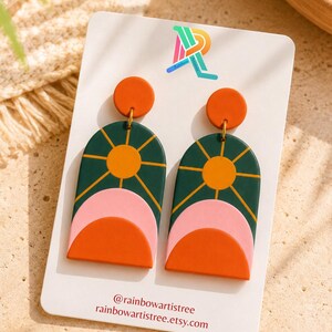 Peut inclure: Une paire de boucles d'oreilles colorées avec un motif de soleil. Les boucles d'oreilles présentent une forme d'arche vert foncé avec un soleil jaune, des demi-cercles roses et oranges. Le texte "@rainbowartistree rainbowartistree.etsy.com" est présent.