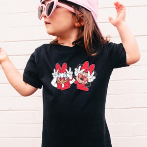 A young girl wearing a black t-shirt with a graphic of two cartoon characters, Minnie Mouse and Dora the Explorer, standing against a white brick wall.