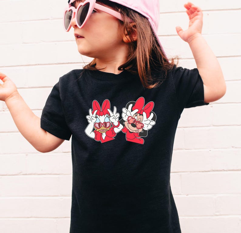 A young girl wearing a black t-shirt with a graphic of two cartoon characters, Minnie Mouse and Dora the Explorer, standing against a white brick wall.