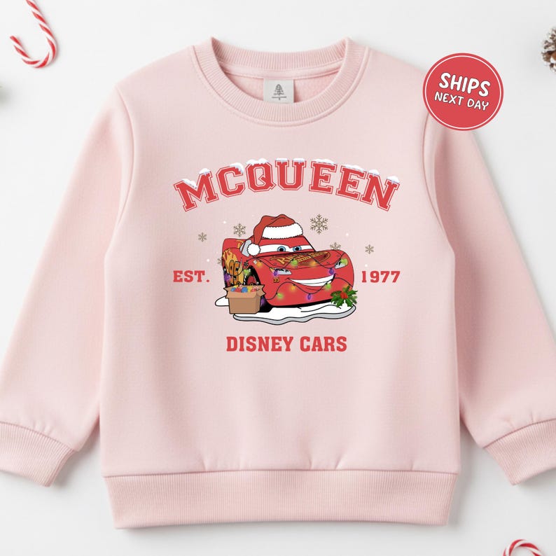 A pink sweatshirt with a graphic design featuring a character from the Disney movie Cars, along with text and a red circular sticker indicating that the sweatshirt is for shipping next day.
