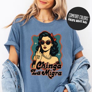 A woman wearing a blue t-shirt with a graphic design featuring a woman with sunglasses and the text "Chinga La Migra" (Spanish for "chilling").