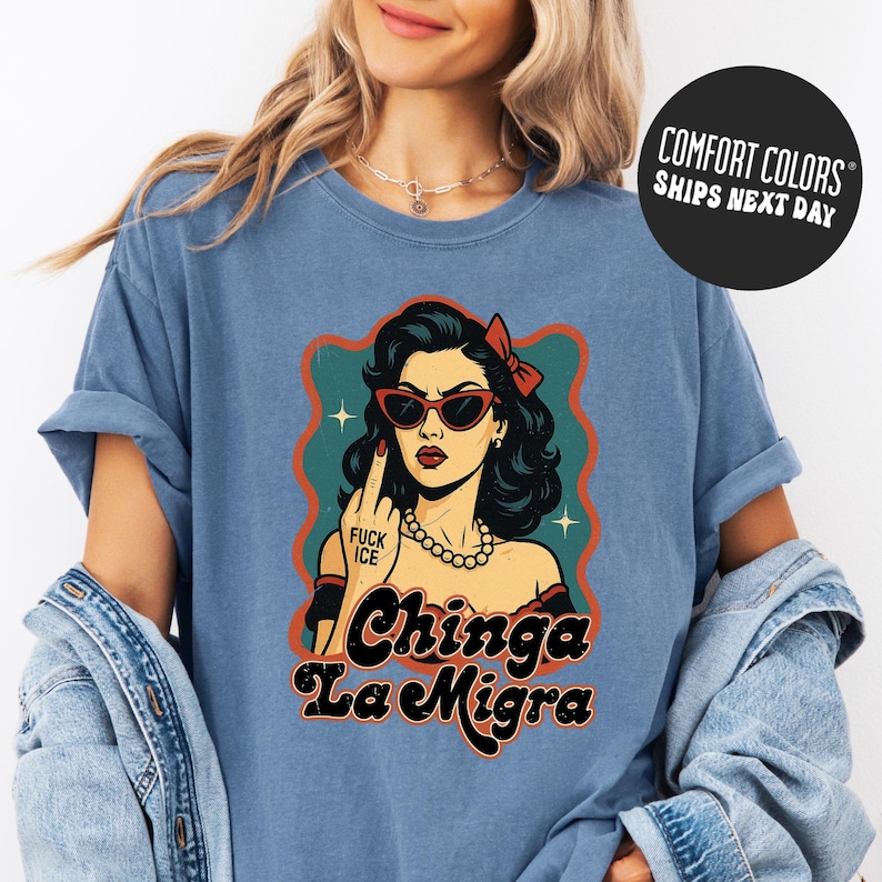 A woman wearing a blue t-shirt with a graphic design featuring a woman with sunglasses and the text "Chinga La Migra" (Spanish for "chilling").
