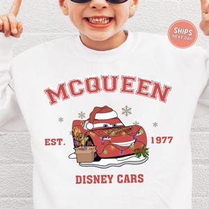 A person wearing a white sweatshirt with a graphic of a red car and a Santa hat, along with the text "McQueen" and "Disney Cars" printed on it.