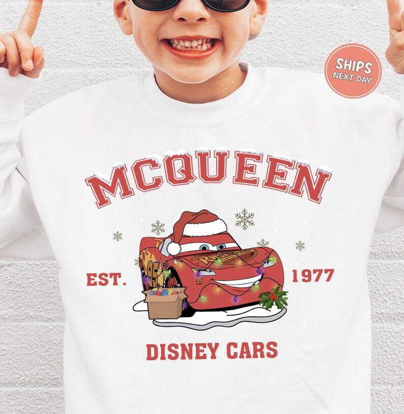 A person wearing a white sweatshirt with a graphic of a red car and a Santa hat, along with the text "McQueen" and "Disney Cars" printed on it.