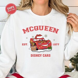 A white sweatshirt with a graphic design featuring a red car and snowflakes, along with the text "McQueen" and "Disney Cars" printed on it.