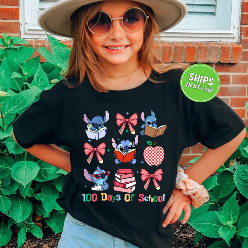 A young girl wearing a black t-shirt with a graphic design of cartoon characters and text, standing in front of a brick wall with green plants.