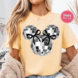 A woman wearing a yellow t-shirt with a graphic of a disco ball and two black bows.